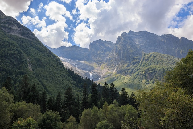 Panoramablick auf die Berge und den weit entfernten Wasserfall im Nationalpark von Dombay, Kaukasus, Russland. Sommerlandschaft, Sonnenscheinwetter und sonniger Tag