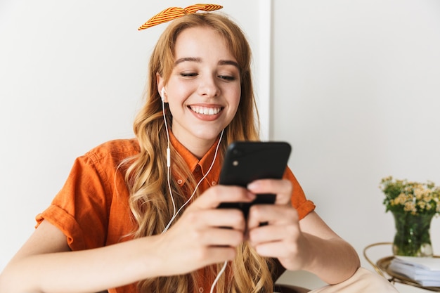 Porträt einer süßen, optimistisch lächelnden jungen blonden Frau zu Hause, die drinnen Musik hört, mit Kopfhörern, die auf dem Stuhl mit Handy sitzen.