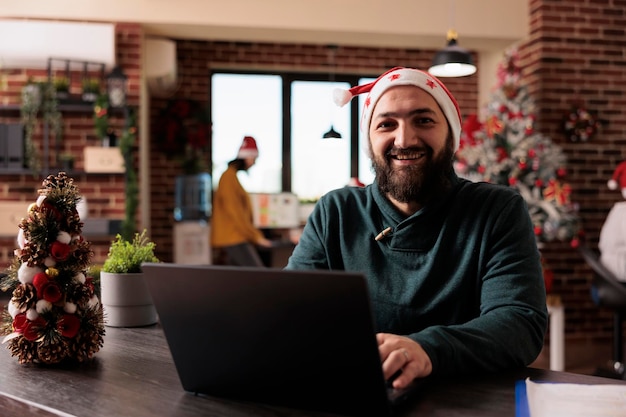 Porträt eines Büroangestellten, der an einem Firmenlaptop arbeitet und im Büro sitzt, das mit Weihnachtsbaum und Lichtern, festlichen Ornamenten geschmückt ist. Lächelnder Mann mit Weihnachtsmütze während der saisonalen Weihnachtszeit.