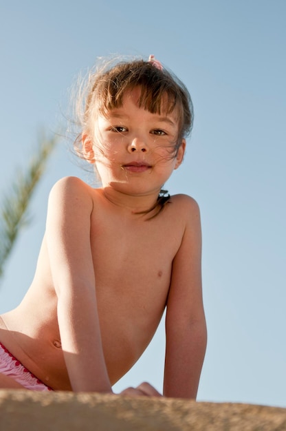 Foto porträt eines hemdlosen mädchens, das auf sand gegen einen klaren himmel sitzt