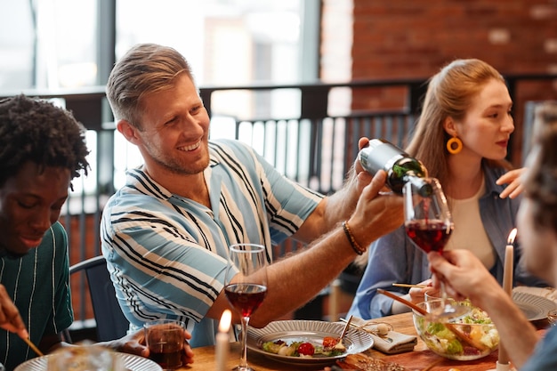 Porträt eines lächelnden Mannes, der Wein in Glas gießt, während er eine Dinnerparty mit Freunden in einem gemütlichen Set genießt