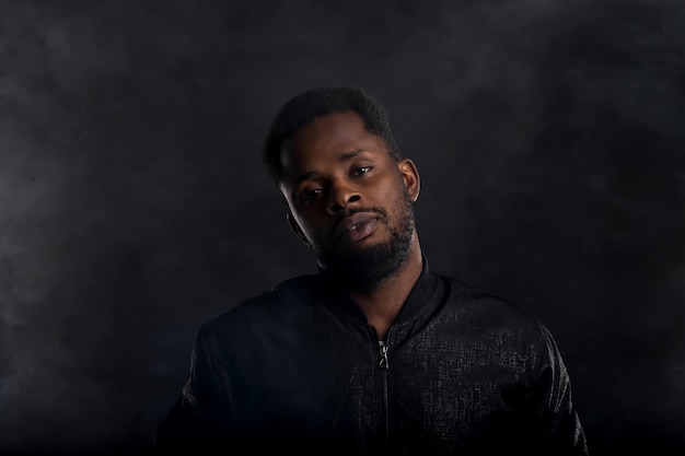 Foto retrato de closeup altamente detalhado de atraente jovem africano vestindo jaqueta preta, olhando para a câmera com uma expressão séria e pensativa em fundo escuro. emoções e sentimentos humanos.