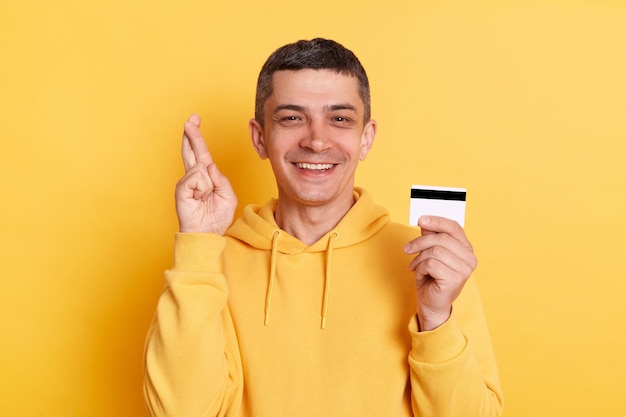 Retrato de homem otimista encantado vestindo capuz estilo casual posando isolado sobre fundo amarelo, segurando o cartão de crédito e cruzando o dedo espera obter dinheiro por conta