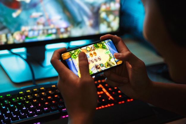 Foto retrato de um menino adolescente jogando videogames em smartphone e computador em uma sala escura usando