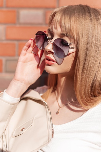 Retrato feminino urbano ensolarado de uma linda jovem com óculos de sol vintage na moda e roupas casuais fica perto de uma parede de tijolos Estilo feminino Moda e beleza