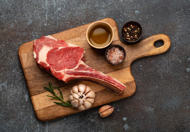 Rohes marmoriertes Tomahawk-Fleischsteak auf Holzbrett mit Kräutern und Gewürzen, konkreter Hintergrund. Premium Rindersteak mit Knochen, bratenfertig, Draufsicht, Steakhaus oder Hausmannskost