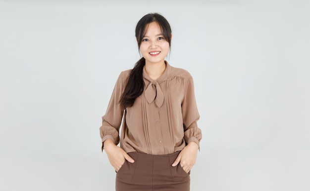 Schönes asiatisches Mädchen lächelt fröhlich als freundliche Emotion und selbstbewusst, während sie mit glücklichem Gesicht und attraktiver charmanter brauner Bluse als hübsches lässiges Outfit für den weiblichen Lebensstil steht.