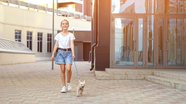 Schulmädchen geht mit dem Hund die Straße entlang. Teenager-Mädchen in weißem T-Shirt und blauen Jeans-Shorts mit Welpen an der Leine geht auf Bürgersteig vorbei an Stadtgebäuden