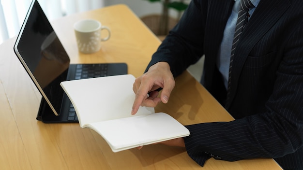 Seitenansicht des Geschäftsmannes, der Informationen auf leerem Notizbuch liest, während mit digitalem Tablett auf dem Tisch gearbeitet wird