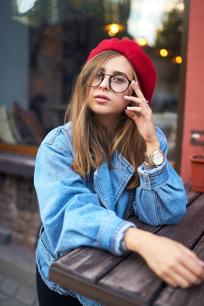 Sommerliches sonniges Lebensstilmodeporträt der jungen stilvollen Hipsterfrau, die auf Straße geht und niedliches trendiges Outfit mit rotem Hut trägt