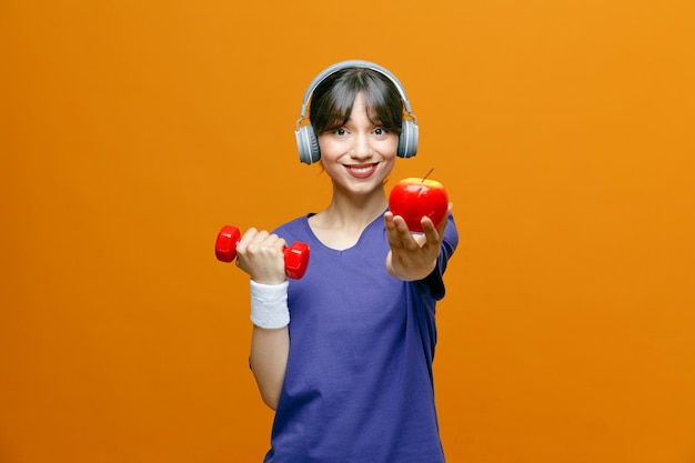 Sportlich schöne Frau in Sportbekleidung mit Kopfhörern auf dem Kopf, die Übungen mit einer Hantel macht, die einen Apfel hält und in die Kamera schaut, glücklich und positiv lächelnd fröhlich über orangefarbenem Hintergrund steht