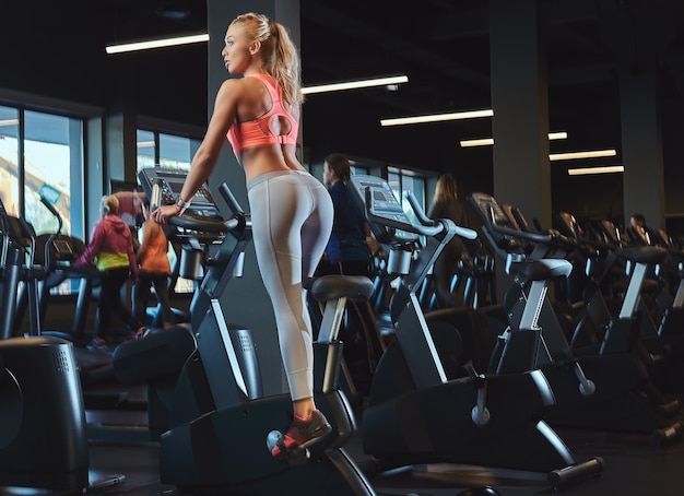 Sportliche blonde Frau, die in einem modernen Fitnesscenter auf einem Heimtrainer trainiert. Fitness-Lifestyle im Sportverein.