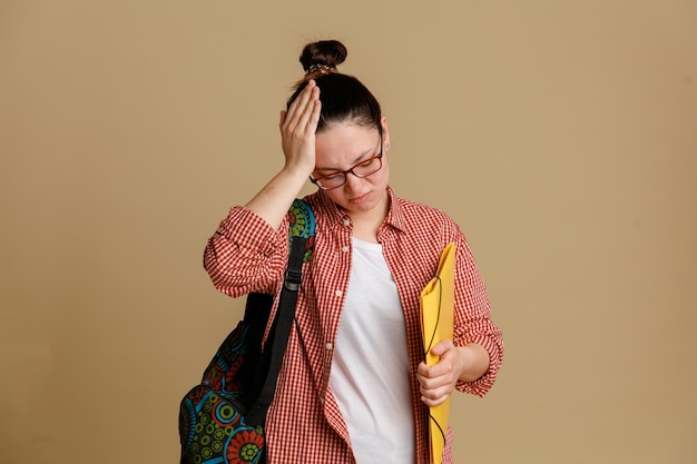 Studentin junge Frau in legerer Kleidung mit Brille und Rucksack mit Ordner, die mit der Hand auf dem Kopf verwirrt aussieht, weil sie einen großen Fehler vor braunem Hintergrund hat