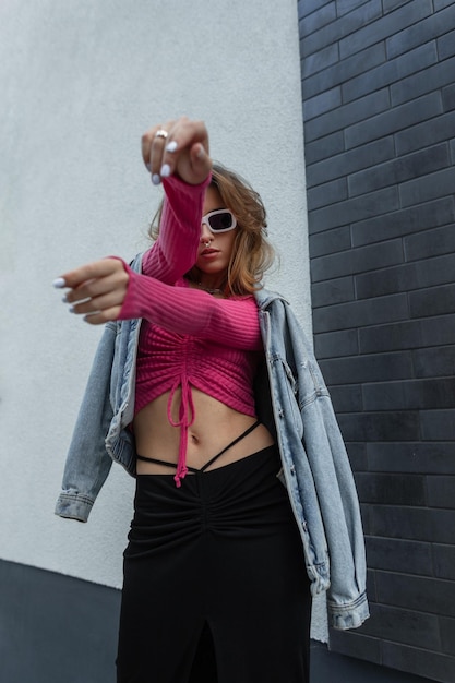 Stylische schöne junge rothaarige Frau Model-Tänzerin mit modischer weißer Sonnenbrille in trendiger urbaner heller Kleidung mit rosa Crop-Top-Jeansjacke und schwarzem langem Rock, die auf der Straße tanzen