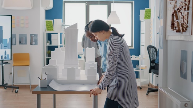 Team aus verschiedenen Architekten, die gemeinsam am Gebäudemodelldesign mit Blaupausen arbeiten. Frauen, die Skizze und Maquette auf dem Tisch verwenden, um das Baulayout und die Immobilienstruktur zu planen.