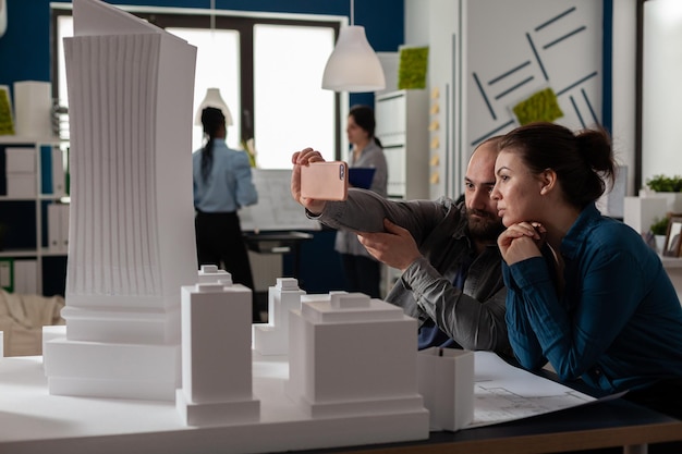 Team von zwei Architektenkollegen, die ein Smartphone in einer Videoanrufkonferenz verwenden, wobei der Kunde einen Schreibtisch mit Gebäudemodellen präsentiert. Architekturingenieure, die mit Smartphone und Maquette zusammenarbeiten.