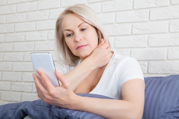 Traurige ältere Frau, die auf Bett mit Smartphone sitzt.