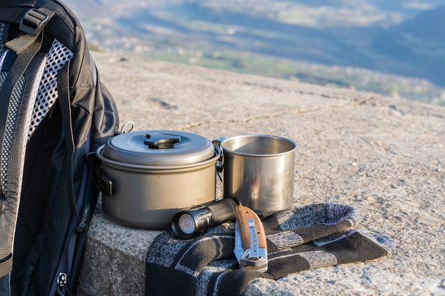 Trekking- oder Wanderausrüstung auf Betonzaun. Rucksack, Socken, Metallbecher, Wasserkocher, Klappmesser und Taschenlampe. Konzept für Outdoor-Aktivitäten.