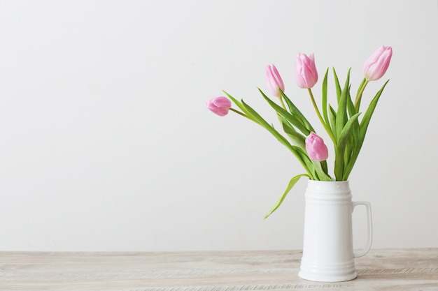 Tulipas cor de rosa no jarro de cerâmico branco na mesa de madeira na parede do fundo branco