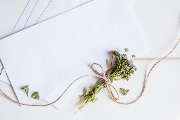 Um envelope branco em branco com um pequeno buquê de flores silvestres amarrado com barbante em uma superfície de madeira branca