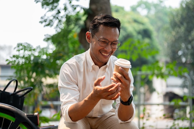 Um feliz empresário asiático senta-se ao ar livre desfrutando de uma pausa para café no parque público em um dia brilhante