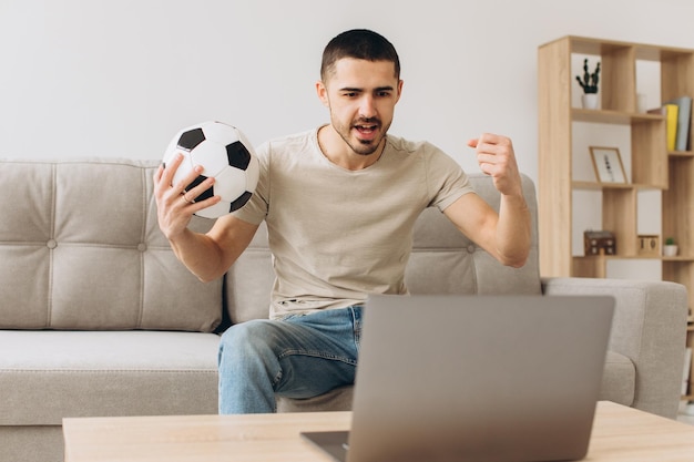 Foto um homem sentado em um sofá na sala segurando uma bola e torcendo pelo time de futebol assistindo a uma partida em um laptop