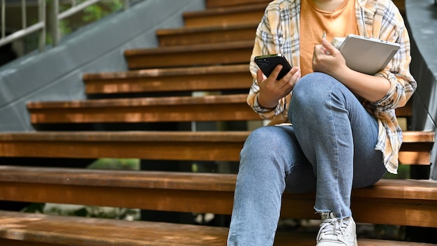 Uma mulher usa seu smartphone enquanto está sentada nas escadas no parque de seu campus imagem recortada