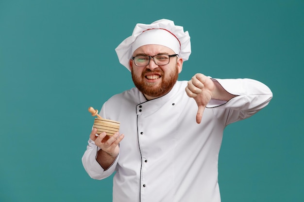 Unzufriedener junger männlicher Koch mit einheitlicher Brille und Mütze, die eine Holzschale mit Honigschöpflöffel darin hält und in die Kamera schaut, die den Daumen nach unten zeigt, isoliert auf blauem Hintergrund