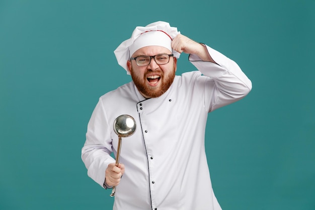 Wütender junger männlicher Koch mit Brille, Uniform und Kappe mit Schöpfkelle, die zeigen, ob Sie dumme Geste sind, die in die Kamera schaut und isoliert auf blauem Hintergrund schreit