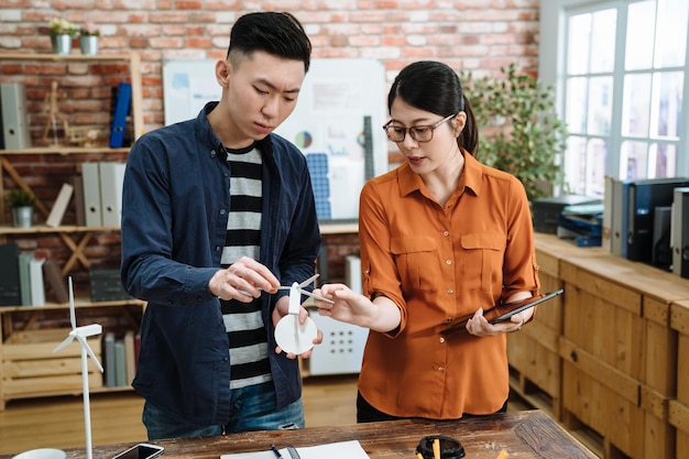 Zwei Architekten asiatischer Japaner, Mann und Frau, die im Büro an einem Bauprojekt arbeiten. junge männliche und weibliche mitarbeiter, die an einem holztisch an einem umweltfreundlichen arbeitsplatz stehen und über eine modellwindmühle diskutieren