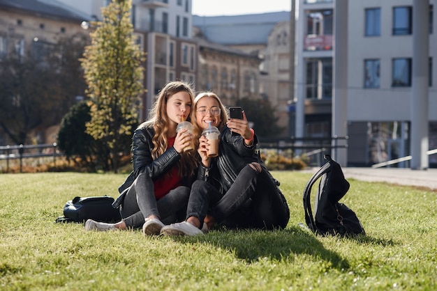 Zwei stilvolle attraktive Teenager-Mädchen mit Smoothie und Smartphone sitzen auf dem Rasen. Freizeit mit besten Freunden.