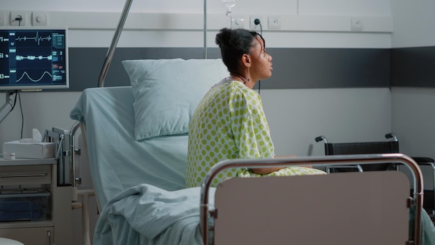 Free photo african american patient with sickness sitting in hospital ward bed, waiting to receive treatment and medical assistance from doctor. young woman with iv drip bag and heart rate monitor