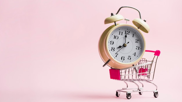 Alarm clock in miniature shopping trolley against pink background