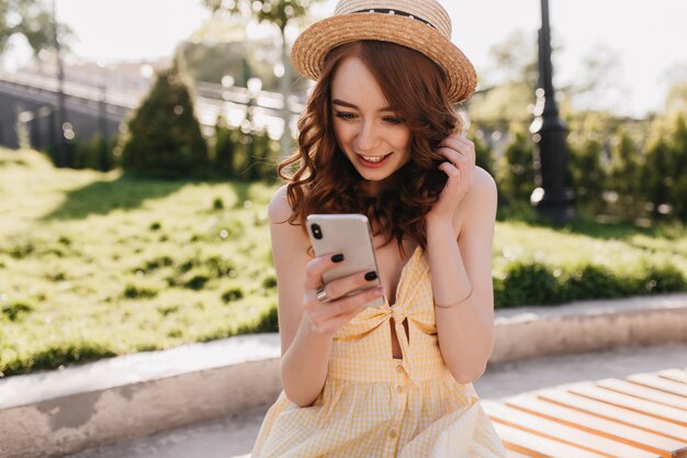 Free photo amazed ginger young woman read phone message in park. outdoor portrait of lovely elegant girl in yellow dress sitting on bench with smartphone.