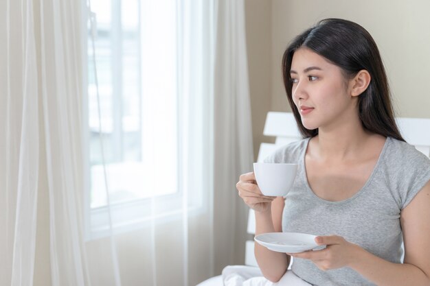 Asian beautiful woman sitting on bed in the bedroom and holding coffee cup in hand 