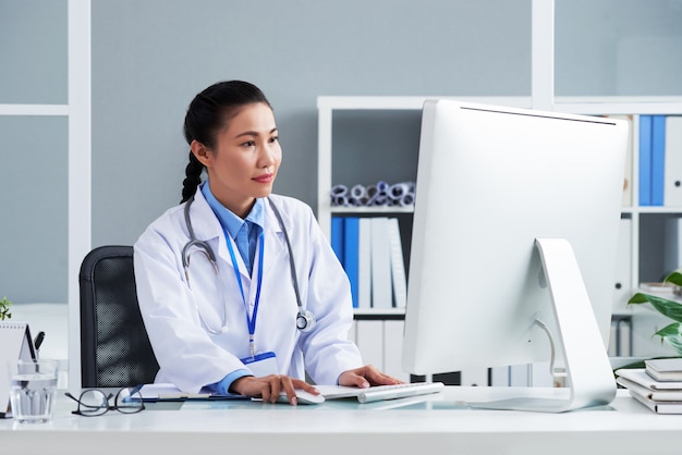 Free photo asian doctor with stethoscope around neck sitting in office and working on computer