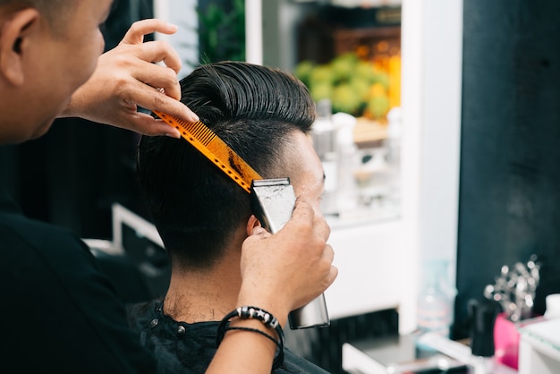 Asian male hairdresser holding comb and trimmer to customer's head