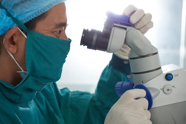 Free photo asian medical researcher in protective apron, mask, hat and gloves peering into microscope