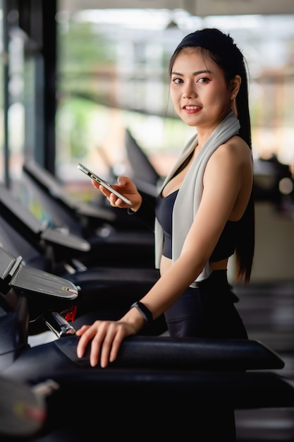 Free photo asian pretty woman wearing sportswear and smartwatch rest on treadmill use smartphone and smartwatch workout app and listening music at modern gym