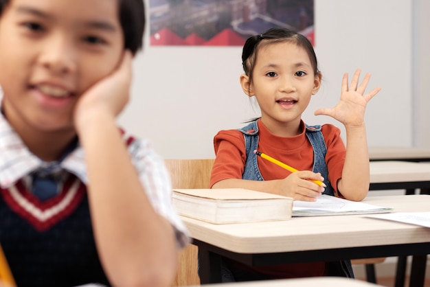 Free photo asian pupils sitting in classroom and girl putting up hand to answer