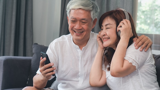 Free Photo asian senior couple relax at home. asian senior chinese grandparents, husband and wife happy wear headphone using mobile phone listen to music while lying on sofa in living room at home concept.