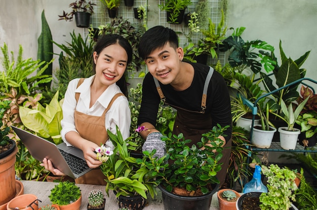 Бесплатное фото Азиатская пара молодых садоводов в фартуке использует садовую технику и портативный компьютер для исследования и ухода за комнатными растениями в теплице