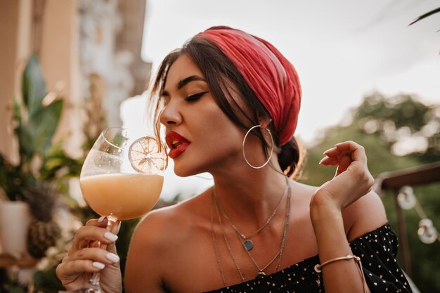 Attractive brunette haired girl with pink headband round silver earrings bright bandana in black shirt drinking cocktail