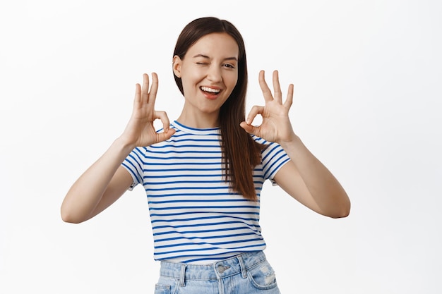 Free Photo not bad, very well. smiling cheerful girl 20s years, winking and showing okay ok sign, encourage you, assure everything under control, all is fine perfect, standing satisfied against white background.