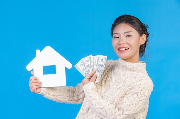 Free photo a beautiful woman wearing a new long sleeved white carpet that holds the house symbol and dollar bills on a blue . trading .