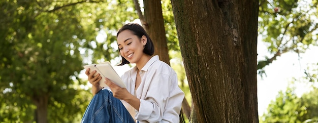 Free photo beautiful young asian girl student sits in park under tree and reading book smiling enjoying warm