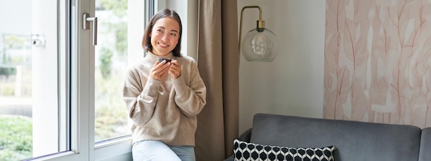 Free photo beautiful young asian woman sitting near window and drinking her coffee holding espresso cup and