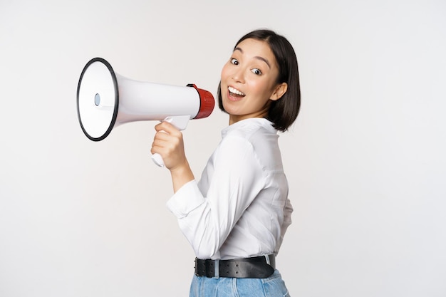 Foto gratuita bella giovane donna asiatica che parla in megafono urla in vivavoce e sorridente facendo l'annuncio gridare informazioni in piedi su sfondo bianco