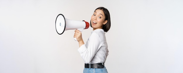 Foto gratuita bella giovane donna asiatica che parla in megafono urla in vivavoce e sorridente facendo l'annuncio gridare informazioni in piedi su sfondo bianco