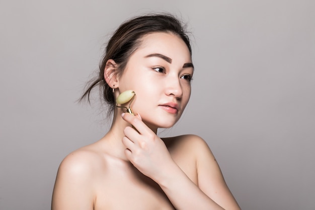 Free Photo beautiful young asian woman using a jade face roller on her flawless skin. beauty face closeup. conceptual of facial treatments with semi precious stones. isolated on gray with copy space